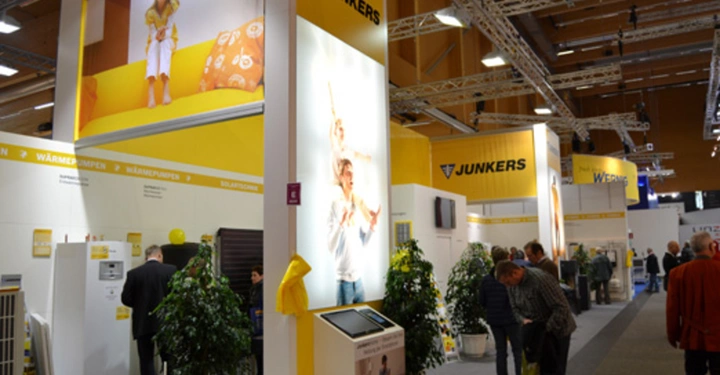 Galerie-Bild 26: Mehrere Menschen betrachten Exponate auf einem Messestand der Firma Junkers in einer Halle mit gelben und weißen Wänden und Pflanzen in Töpfen. Oben sind Werbebilder und Text: WÄRMEPUMPEN, JUNKERS.