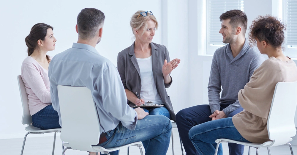 Mediatorin sitzt im Kreis mit vier weiteren Gesprächsteilnehmern auf weißen Sesseln in weißem Raum. Es wird diskutiert.