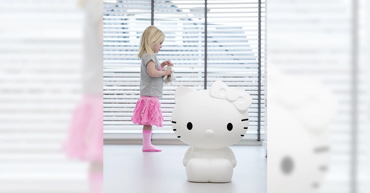 Galerie-Bild 4: Weiße Hello Kitty-Figur sitzt auf dem Boden. Ein kleines Mädchen im Hintergrund hält ein Kuscheltier in ihren Händen. Lichtdurchfluteter Raum mit großen Fensterjalousien im Hintergrund.