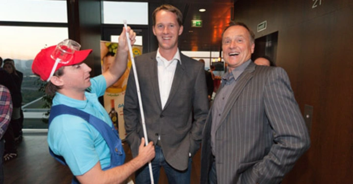 Galerie-Bild 50: Ein Mann in einem blauen Shirt und roter Kappe misst mit einem Maßband die Höhe eines Mannes in einem grauen Anzug im Inneren eines modernen Gebäudes während ein lachender Mann daneben steht.