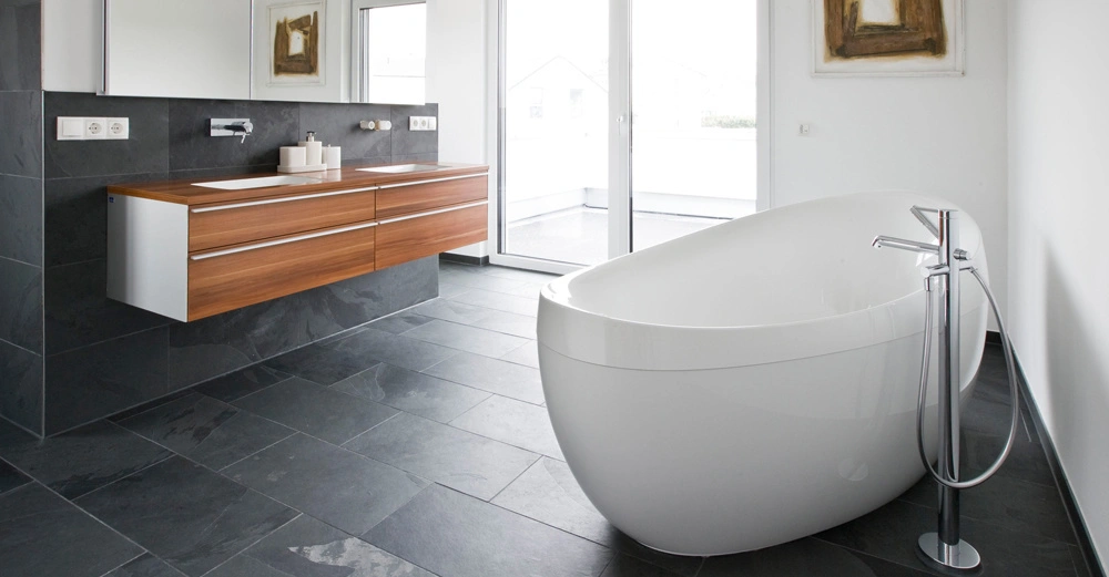 Freistehende weiße Badewanne steht auf dunkelgrauem Steinboden vor großer Glasschiebetür; daneben brauner Holzwaschtisch mit weißen Waschbecken und Spiegel. Wandkunst über der Badewanne.