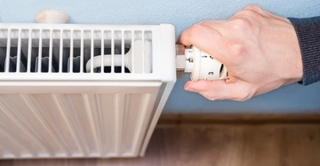 Eine Hand dreht den Thermostat eines weißen Heizkörpers in einem Raum mit hellblauer Wand und Holzboden