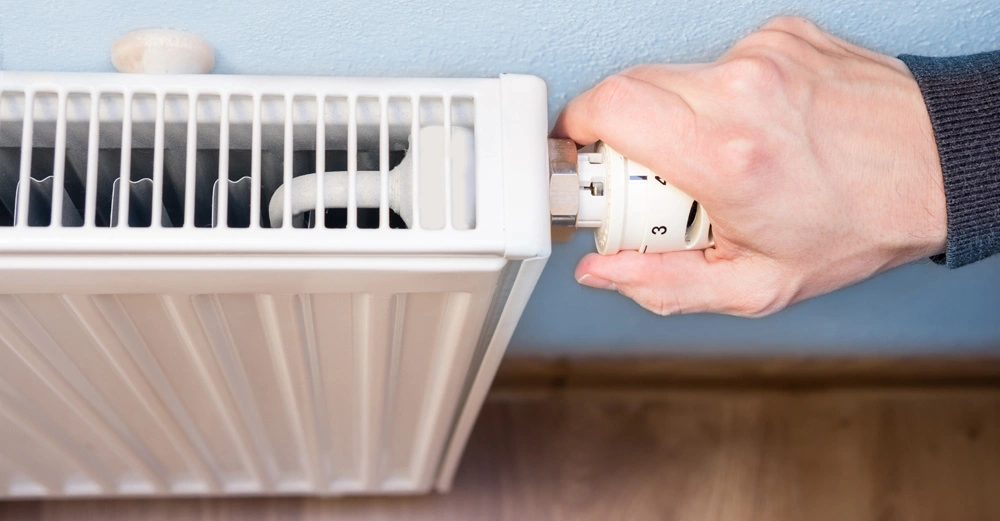 Eine Hand dreht den Thermostat eines weißen Heizkörpers in einem Raum mit hellblauer Wand und Holzboden