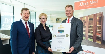 Drei Personen halten ein großes Zertifikat in einem modernen Büro mit Glasfenstern und Werbebanner im Hintergrund. Text auf dem Zertifikat: QOA Zertifikat Delfin Wellness Austria Gütezeichen Österreichischer Musterbetrieb