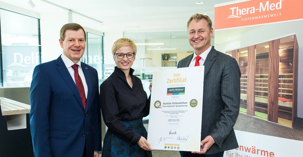 Drei Personen halten ein großes Zertifikat in einem modernen Büro mit Glasfenstern und Werbebanner im Hintergrund. Text auf dem Zertifikat: QOA Zertifikat Delfin Wellness Austria Gütezeichen Österreichischer Musterbetrieb
