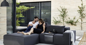 Zwei Menschen sitzen auf einem modernen grauen Sofa draußen Sie lachen und umarmen sich Der Hof hat Kiesboden einige Pflanzen und eine helle Wand im Hintergrund