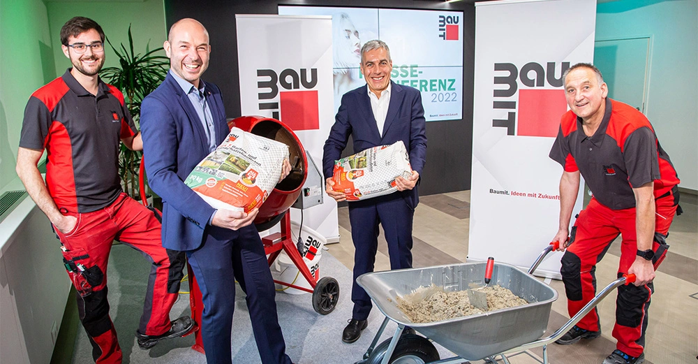 Fünf Männer stehen in einem hellen Raum, zwei tragen Anzüge und halten Zementsäcke, drei tragen Arbeiterkleidung, daneben eine Mischmaschine und Schubkarre mit Sand. Text: Baumit Pressekonferenz 2022 Baumit Ideen mit Zukunft