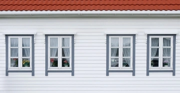 Vier weiße Fenster mit Blumen dahinter befinden sich an einer weißen Holzhauswand unter einem roten Ziegeldach.