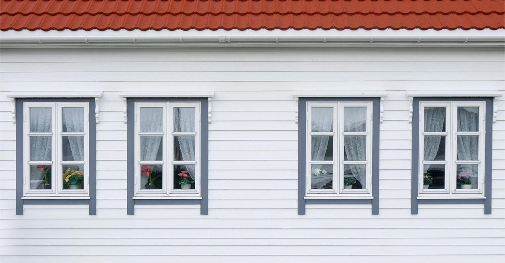 Vier weiße Fenster mit Blumen dahinter befinden sich an einer weißen Holzhauswand unter einem roten Ziegeldach.