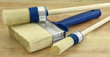 zwei Stupfpinsel unterschiedlicher Größe und ein breiter Flachpinsel leigen nebeneinander auf hellem Holztisch. Pinsel haben hölzerne bzw. blau lackierte Griffe.