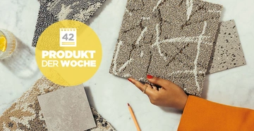 Eine Hand mit rotem Nagellack hält eine Textilprobe auf einem Marmortisch neben Musterkarten und einem Wasserglas mit Zitrone. Text: 42 PRODUKT DER WOCHE