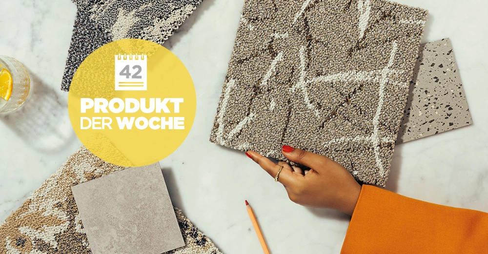 Eine Hand mit rotem Nagellack hält eine Textilprobe auf einem Marmortisch neben Musterkarten und einem Wasserglas mit Zitrone. Text: 42 PRODUKT DER WOCHE