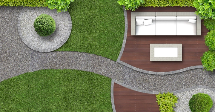 Weiße Couchgarnitur mit Kissen auf einer hölzernen Terrasse neben einem weißem rechteckigen Tisch umgeben von grünem Rasen und einem kurvigen Steinweg mit Bäumen und Pflanzen.