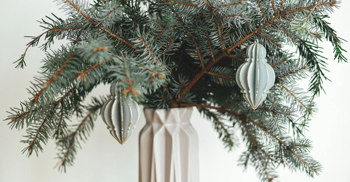 Vase hält Tannenzweige mit zwei hängenden silbernen Weihnachtsornamenten in einem hellen Innenraum