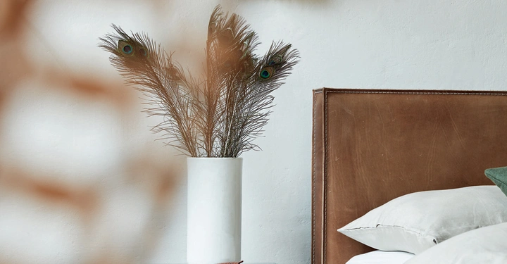 Weiße Vase mit Pfauenfedern auf einem dunklen Nachttisch neben einem Bett in einem Schlafzimmer mit Holzfußboden und dekorativen Elementen an der Wand