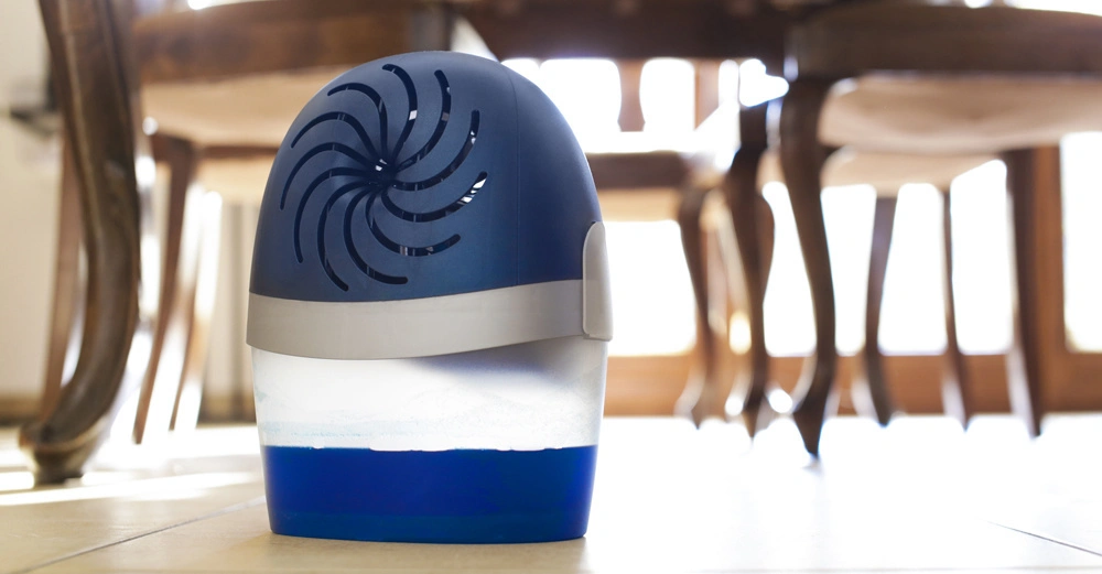 Ein blauer Luftentfeuchter steht auf einem gefliesten Boden und arbeitet in einem hell erleuchteten Esszimmer mit Holzmöbeln im Hintergrund.