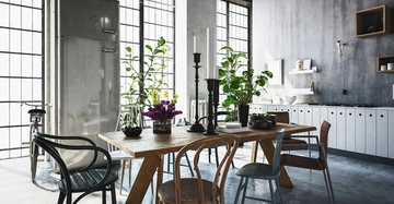 Großer Kühlschrank in moderner, industrieller Küche steht, umgeben von einem hölzernen Esstisch mit Stühlen und Pflanzen; hohe Fenster lassen viel Licht hinein