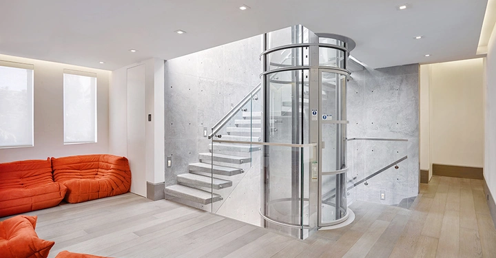 Ein runder, gläserner Aufzug steht still in einem modern eingerichteten Wohnzimmer mit hölzernem Boden, zwei Fenstern und roten Sofas; daneben befindet sich eine Treppe mit Glasgeländer.