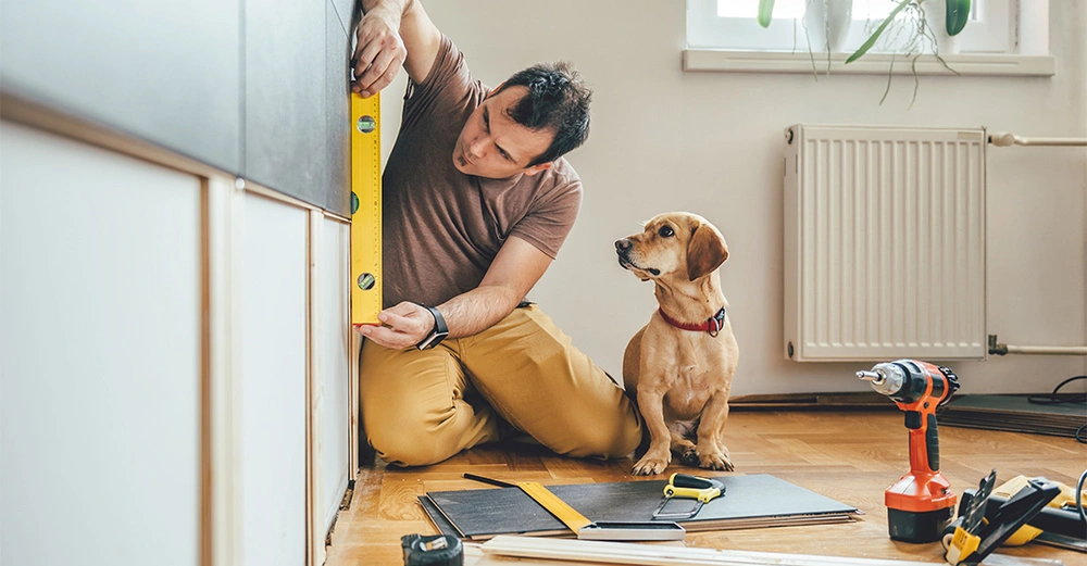 Brünetter Mann misst eine vertikale Oberfläche mit Wasserwaage, sein neugieriger Hund sieht dabei zu.