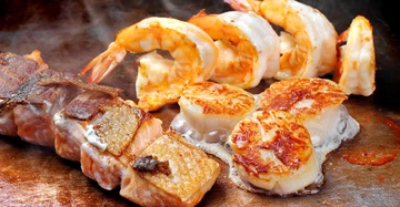 Garnelen Jakobsmuscheln und Lachsfilets brutzeln auf einem heißen Grill in einer Küche die für das Kochen vorbereitet ist.