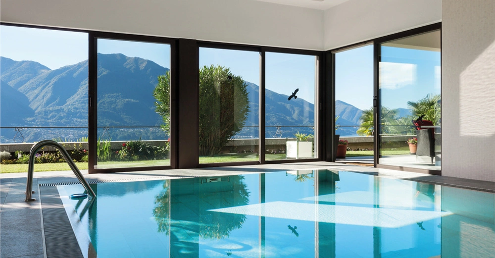 großer Indoorpool mit Ganzglaswänden, die sich öffnen lassen und den Blick in den Garten draußen freigeben. Am Rand stehen gemütliche Liegen. 