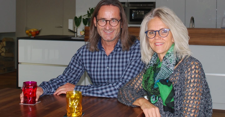 Zwei Personen sitzen an einem Holztisch in einer modernen Küche und lächeln in die Kamera. Einer hält ein rotes Glas, der andere ein goldenes. Obstschale und Pflanzen im Hintergrund.