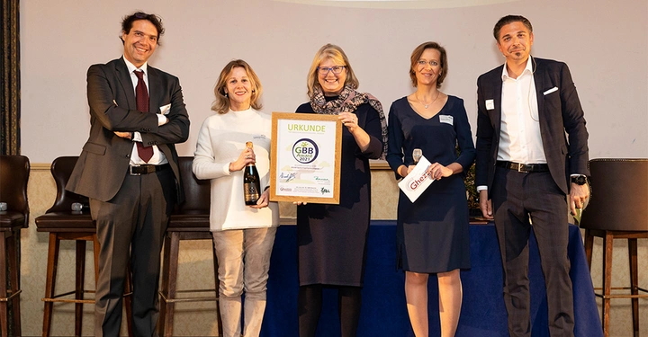 Galerie-Bild 6: Fünf Personen posieren auf einer Bühne eine hält eine Urkunde und eine andere eine Sektflasche. Im Hintergrund sind leere Stühle. Text auf der Urkunde: URKUNDE GBB Award 2021 Text auf der Karte hinten links: Ghezzo