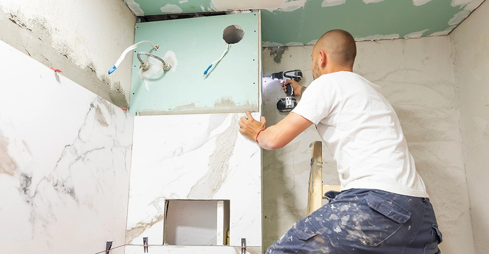 Installateur in grauer Arbeitshose und weißem Shirt steht auf Leiter und bohrt eine Seite der Vorwandinstallation im Badezimmer fest. Bad noch im Trocken-Rohbau