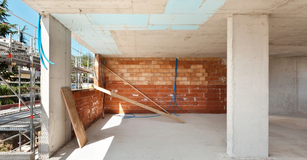 Unvollendete Betonstruktur mit zwei Betonstützen Wände aus roten Ziegelsteinen und blauem Elektrikkabel Baumaterialien stützend an der Wand Außenumgebung zeigt Gerüste und Vegetation unter klarem Himmel.