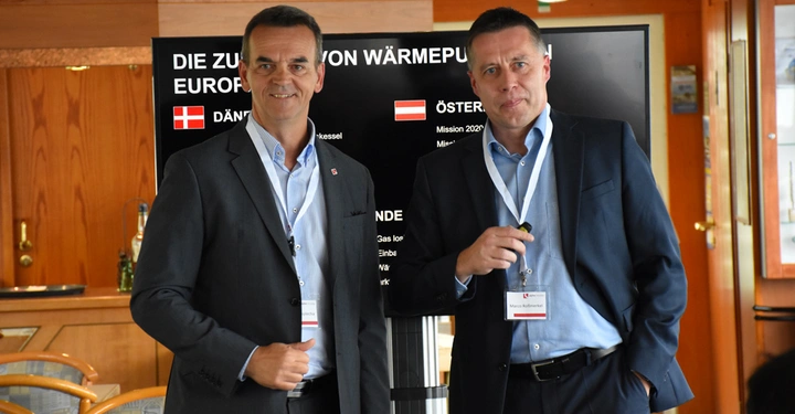 Zwei Männer tragen Anzug und Namensschilder und stehen vor einem großen Bildschirm umgeben von Holzmöbeln. Der Bildschirm zeigt die folgenden Texte: DIE ZUKUNFT VON WÄRMEPUMPEN IN EUROPA DÄN Tysklandkessel ÖSTERREICH Mission 2022 Mis.