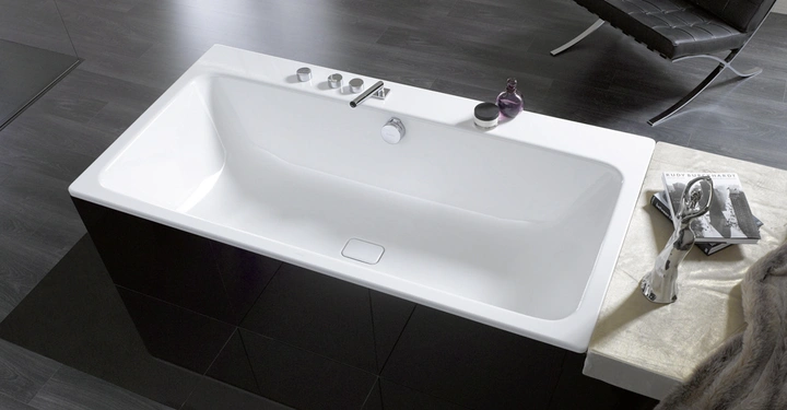 Weiße Badewanne eingebaut in schwarze Fliesen mit moderner Armatur neben einem schwarzen Stuhl in einem Badezimmer mit dunklem Holzboden und einem Tisch, auf dem Bücher und eine Skulptur liegen