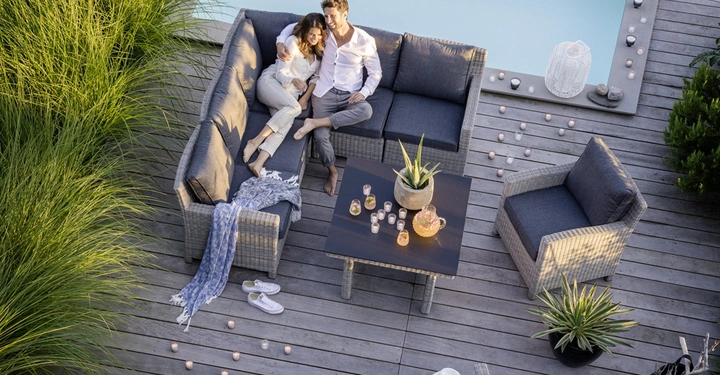 Galerie-Bild 4: Ein Paar sitzt auf einer grauen Couch und entspannt am Pool auf einer Holzterrasse Umgeben von Pflanzen Kerzen und Dekorationen.