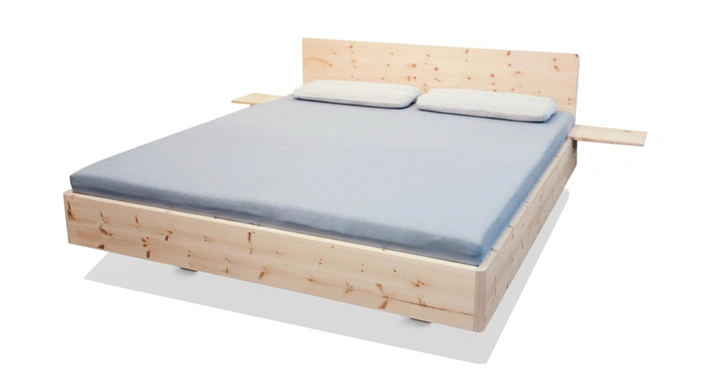 Ein Holz-Doppelbett mit zwei Kopfkissen liegt mittig in einem hell beleuchteten Raum auf weißem Hintergrund