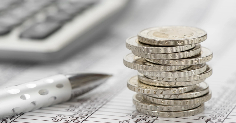 Ein Stapel Münzen steht auf einem Dokument mit Tabellen. Rechts daneben liegt ein silberner Stift und im Hintergrund ist ein unscharfer Taschenrechner zu sehen.