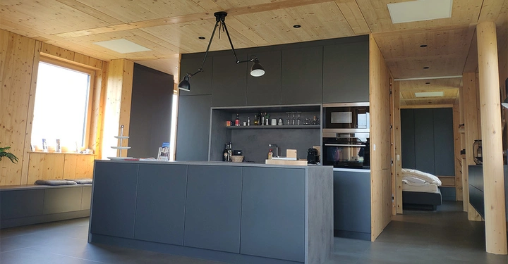 Kücheneinrichtung bestehend aus grauen Schränken und einem Inseltresen mit Hängelampen in einem Raum mit Holzwänden und -decke. Eine Tür führt zu einem Schlafzimmer.
