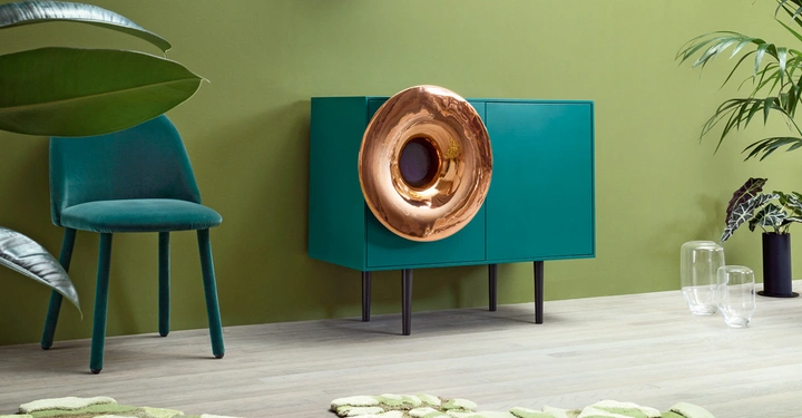 Ein grüner Stuhl steht neben einem türkisfarbenen Schrank mit einem großen kupfernen Blasinstrument darauf in einem modern eingerichteten Raum mit grüner Wand und Pflanzen.