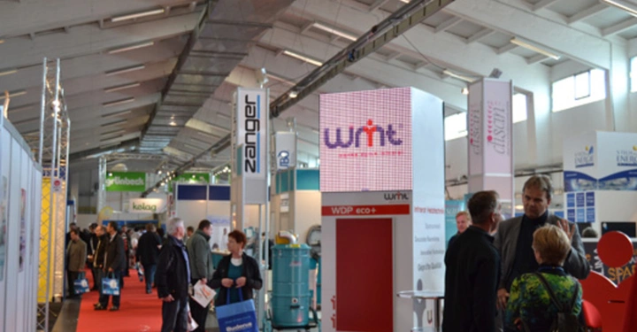 Galerie-Bild 8: Personen gehen und unterhalten sich in einer Messehalle mit roten Teppichen wobei verschiedene Stände und Firmenlogos wie Zanger und wnt-Technik sichtbar sind. Ein heller Raum mit industriellem Design.