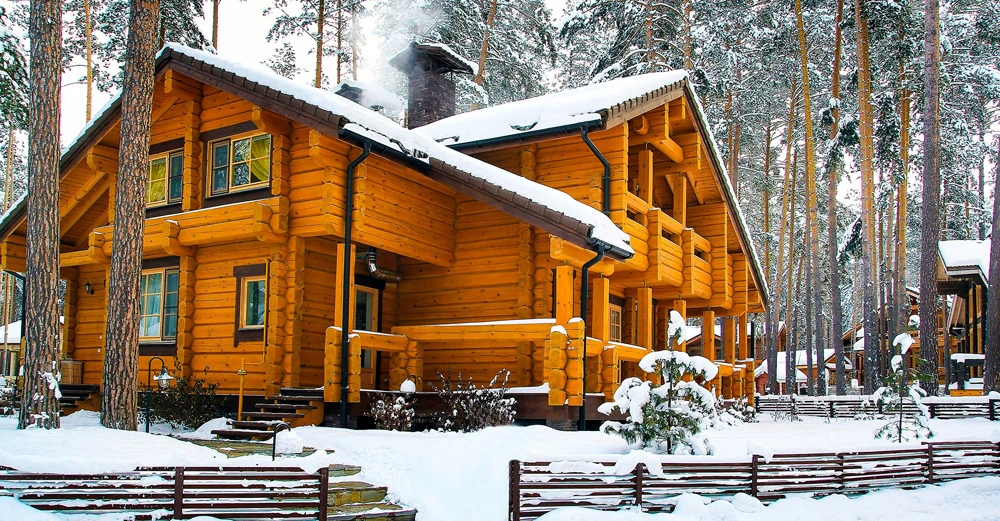 Holzblockhaus mit Rauch aus dem Schornstein steht im verschneiten Waldumfeld mit schneebedecktem Dach Bäumen im Hintergrund und einem Holzlattenzaun im Vordergrund