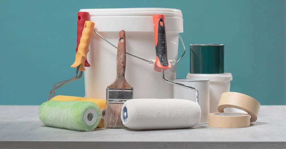 Ein Farbeimer mit Pinseln, Rollen und Krepp vor einem türkisen Hintergrund.
