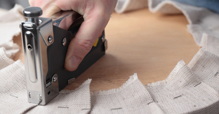 Eine Hand hält einen Tacker und befestigt Stoff auf einer runden Holzfläche in einem handwerklichen Kontext. Der Stoff ist unregelmäßig zugeschnitten und bereits teils festgetackert.