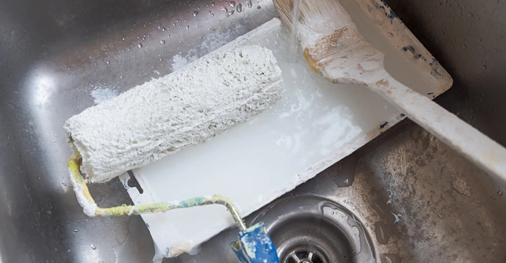 Malerrolle mit blauem Stiehl und Malerpinsel mit weißer Farbe liegen in Edelstahl-Waschbecken und Wasser läuft auf die Pinsel. Sie werden gereinigt. Aufnahme von oben.