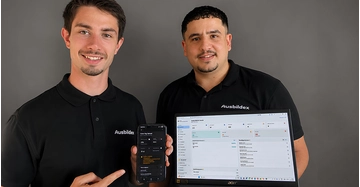 zwei junge Männer in schwarzen Polohemden mit der Aufschrift "Ausbildex" zeigen Handy und Laptop mit Software