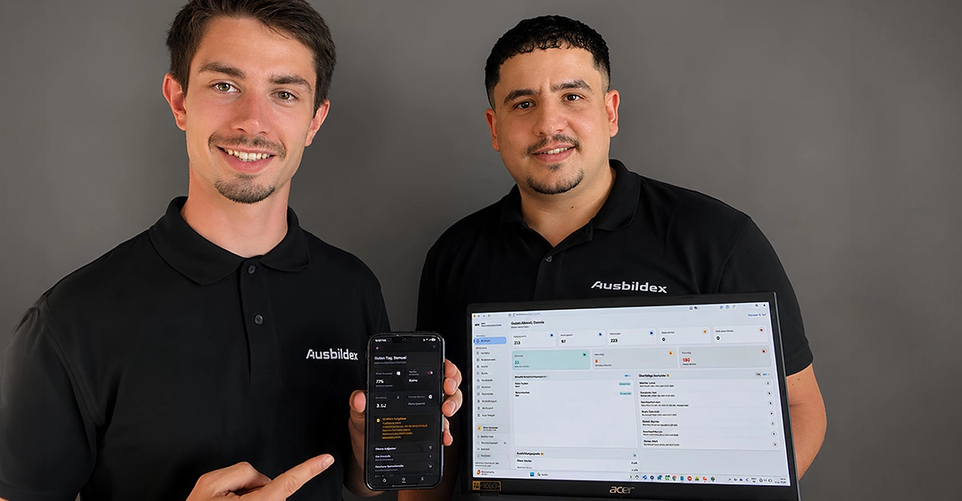zwei junge Männer in schwarzen Polohemden mit der Aufschrift "Ausbildex" zeigen Handy und Laptop mit Software