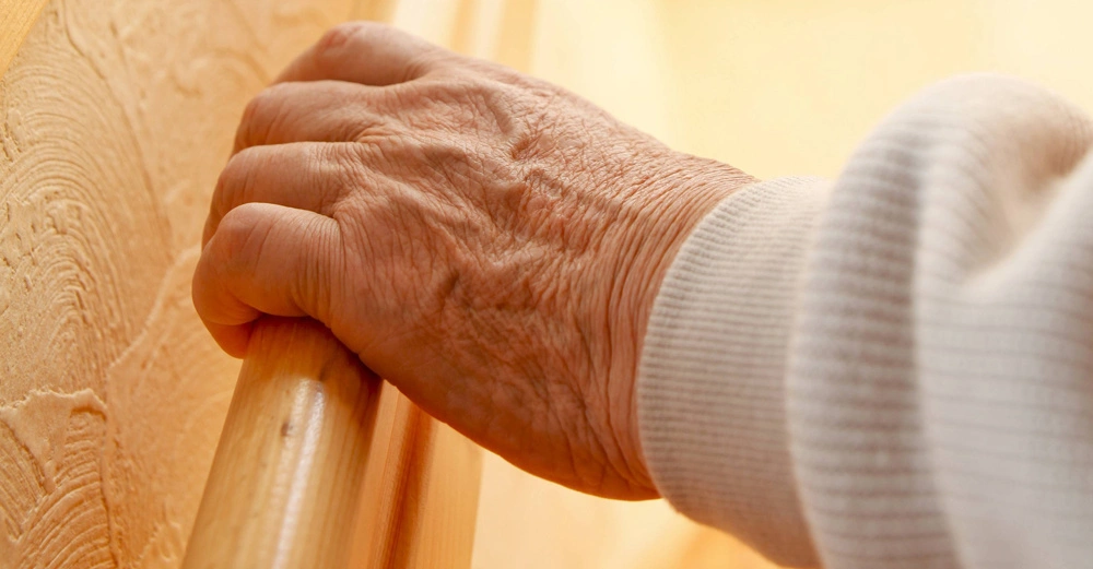 Hand greift ein Geländer in einem gut beleuchteten, holzgetäfelten Innenraum.