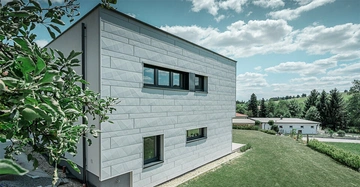 Ein modernes weißes Haus mit rechteckigen Fenstern steht in einer grünen ländlichen Umgebung mit Bäumen, Gras und einem klaren blauen Himmel mit Wolken. Ein Baum befindet sich im Vordergrund links.