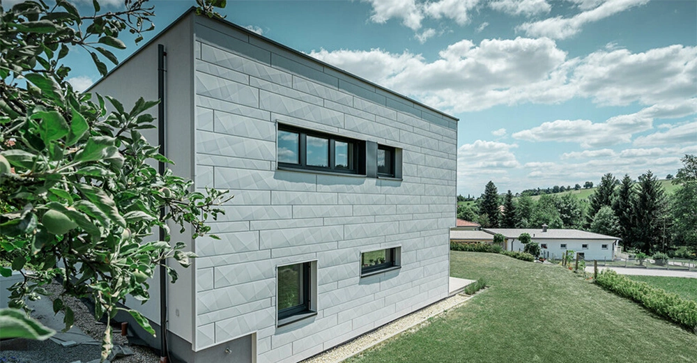 Ein modernes weißes Haus mit rechteckigen Fenstern steht in einer grünen ländlichen Umgebung mit Bäumen, Gras und einem klaren blauen Himmel mit Wolken. Ein Baum befindet sich im Vordergrund links.