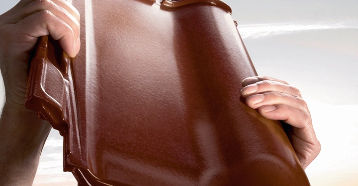 Hände halten eine glänzende rote Dachziegel in die Höhe vor einem Hintergrund aus blauem Himmel und grünen Hügeln