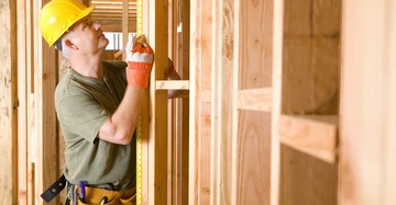 Handwerker mit gelbem Schutzhelm und Handschuhen misst Holzbalken mit Maßband in einem im Bau befindlichen Holzrahmenhaus. Er trägt einen Werkzeuggürtel und befindet sich in einem unvollendeten Raum.
