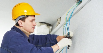 Ein Arbeiter mit gelbem Schutzhelm und Handschuhen zieht Kabel durch ein Loch in der Wand. Der Arbeiter befindet sich in einem Innenraum und ist in Arbeitskleidung gekleidet.