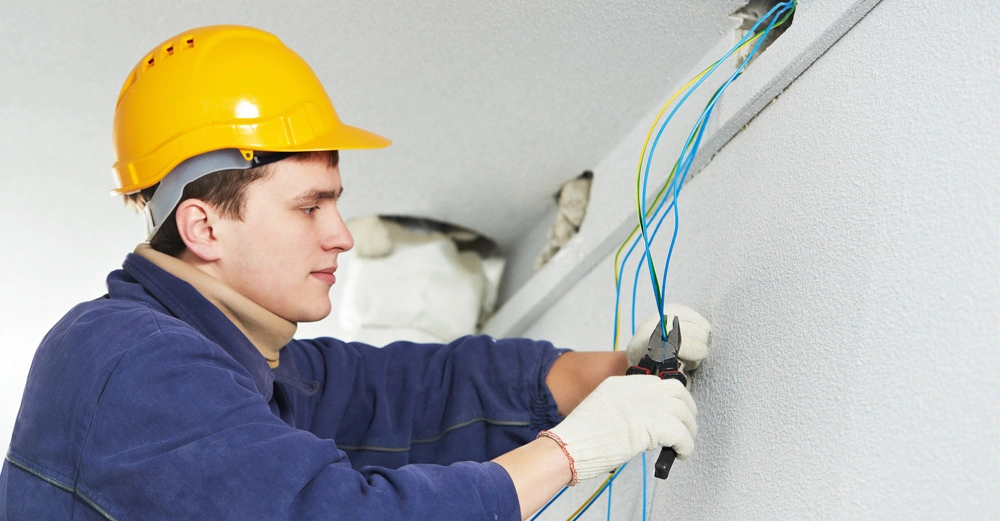 Ein Arbeiter mit gelbem Schutzhelm und Handschuhen zieht Kabel durch ein Loch in der Wand. Der Arbeiter befindet sich in einem Innenraum und ist in Arbeitskleidung gekleidet.